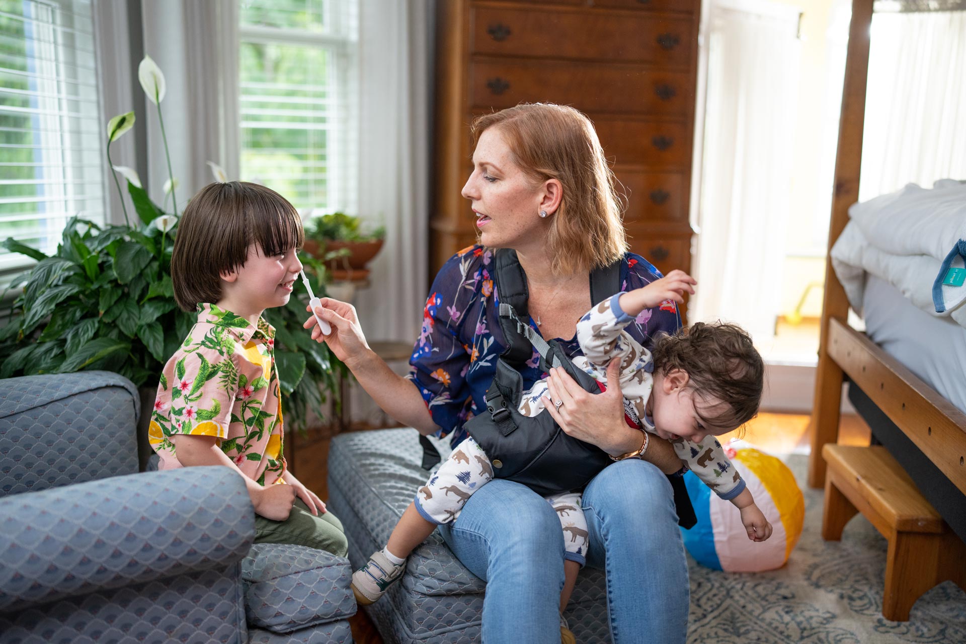 A mother swabs her child's nose while a baby squirms on her lap.