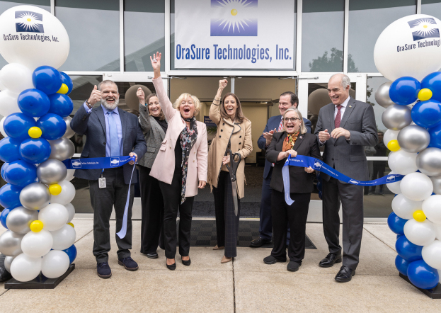 A group of people stand in front of the OraSure Technologies, Inc building. They are cheering and holding a cut ceremonial ribbon and are surrounded by balloons.