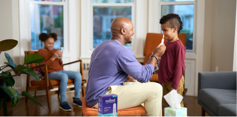 A man swabs a child's nose at home. They are both smiling. Another child is smiling while on their phone in the background.