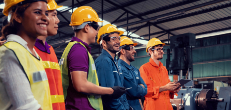 A group of workers stand in a factory wearing hard-hats and safety gear while laughing.