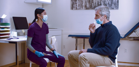 A woman dressed in scrubs sits in a medical exam room and watches a man swab his nose.